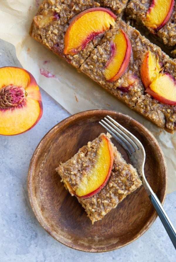 Slice of peach baked oatmeal on a wooden plate with more slices sitting on a piece of parchment paper and half of a fresh peach to the side.