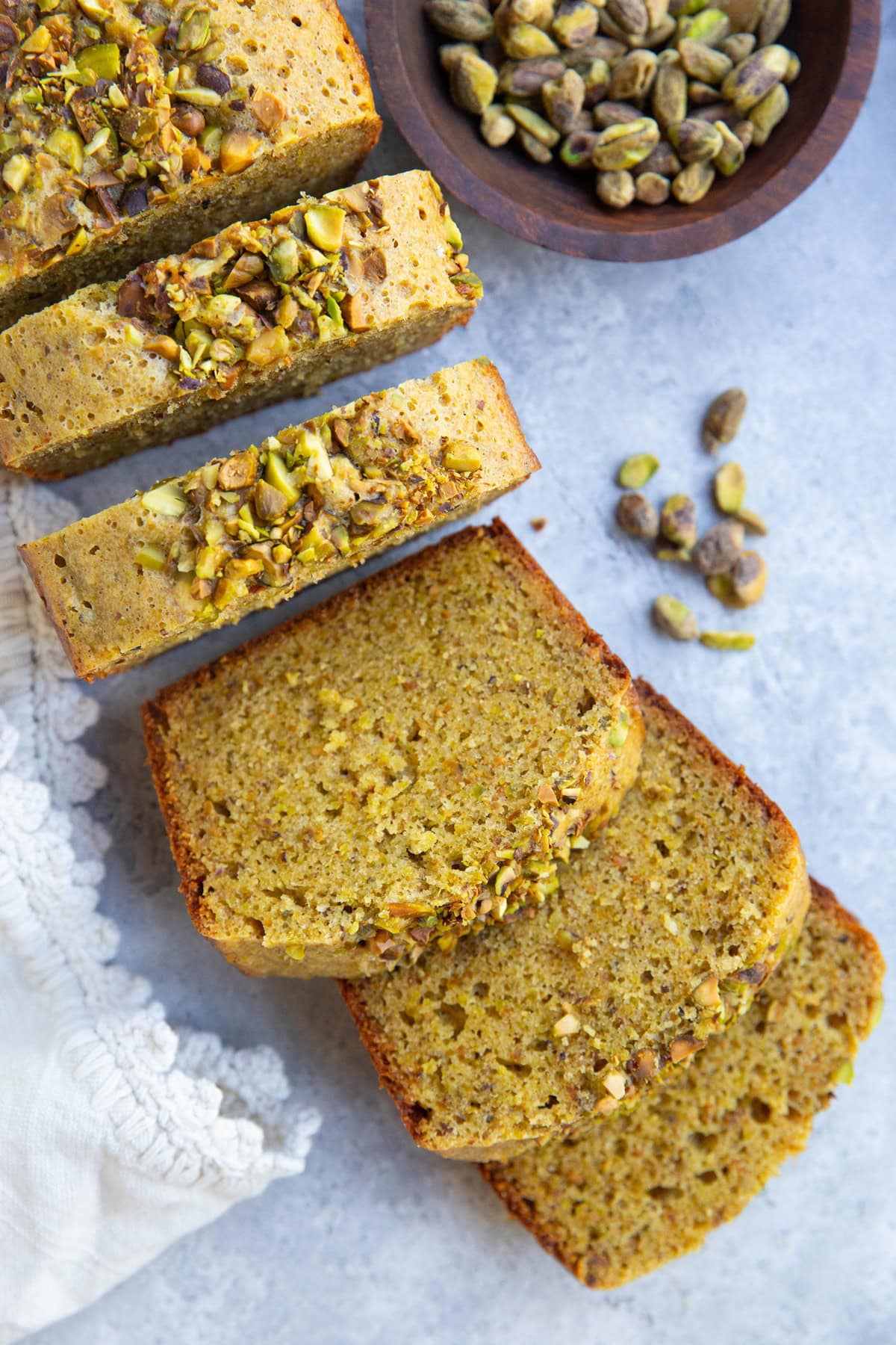 Pistachio cake cut into thick slices with a bowl of raw pistachios to the side and a white napkin.