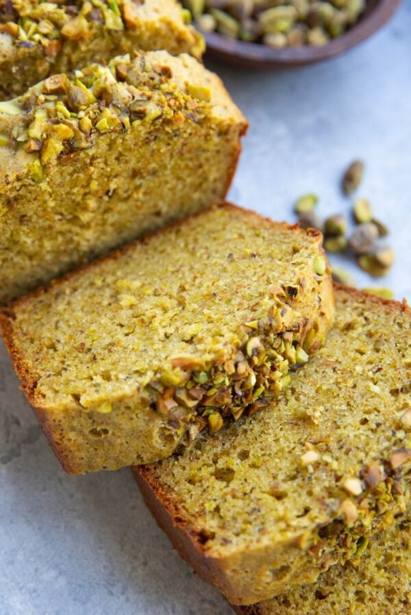Thick slices of pistachio cake with raw pistachios in the background. The slices are in the form of a loaf.