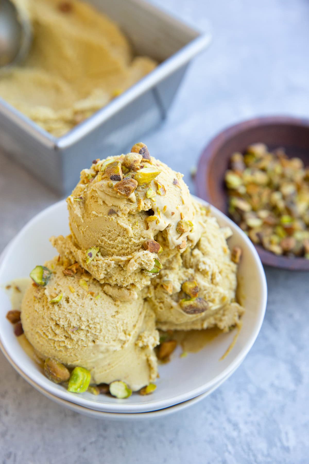 Three scoops of pistachio ice cream in a small bowl, sprinkled with chopped pistachios. A bowl of pistachios and the rest of the ice cream in the background.