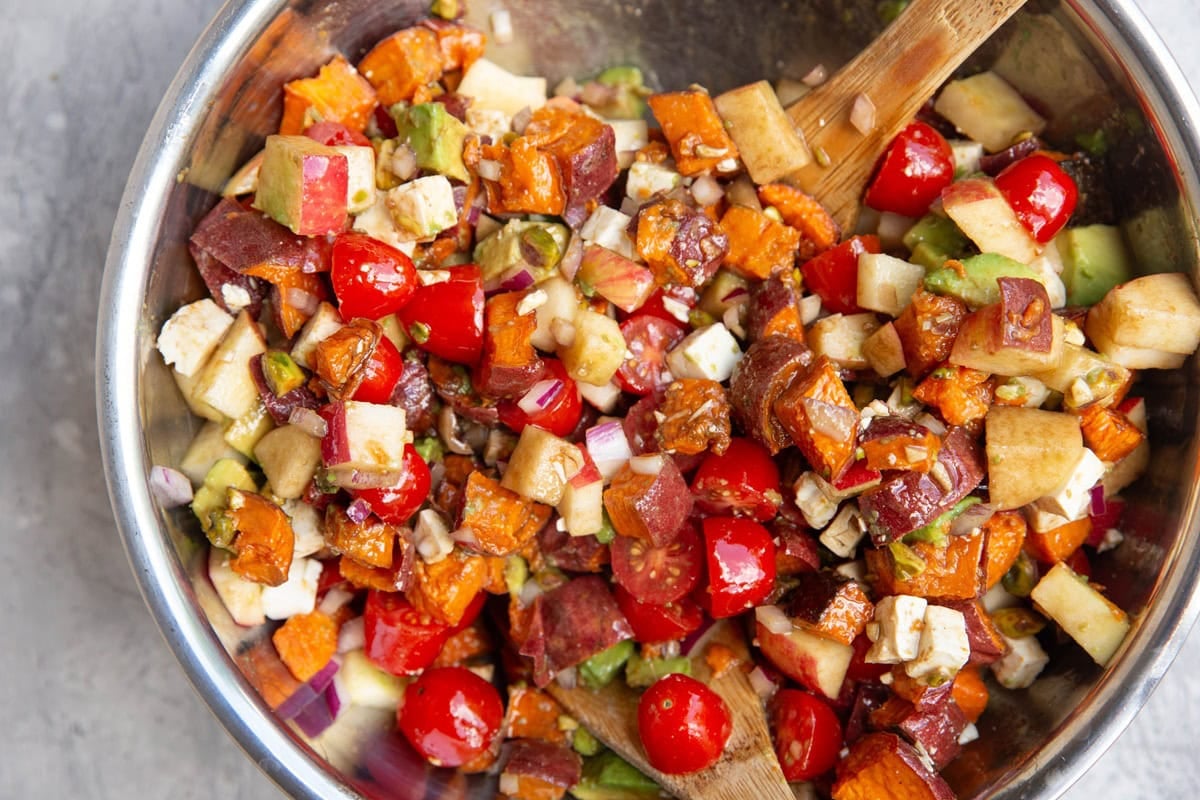 Stainless steel bowl full of roasted sweet potatoes, cherry tomatoes, apples, pistachios, avocado, and more. two wooden spoons are inside the bowl for stirring and serving.
