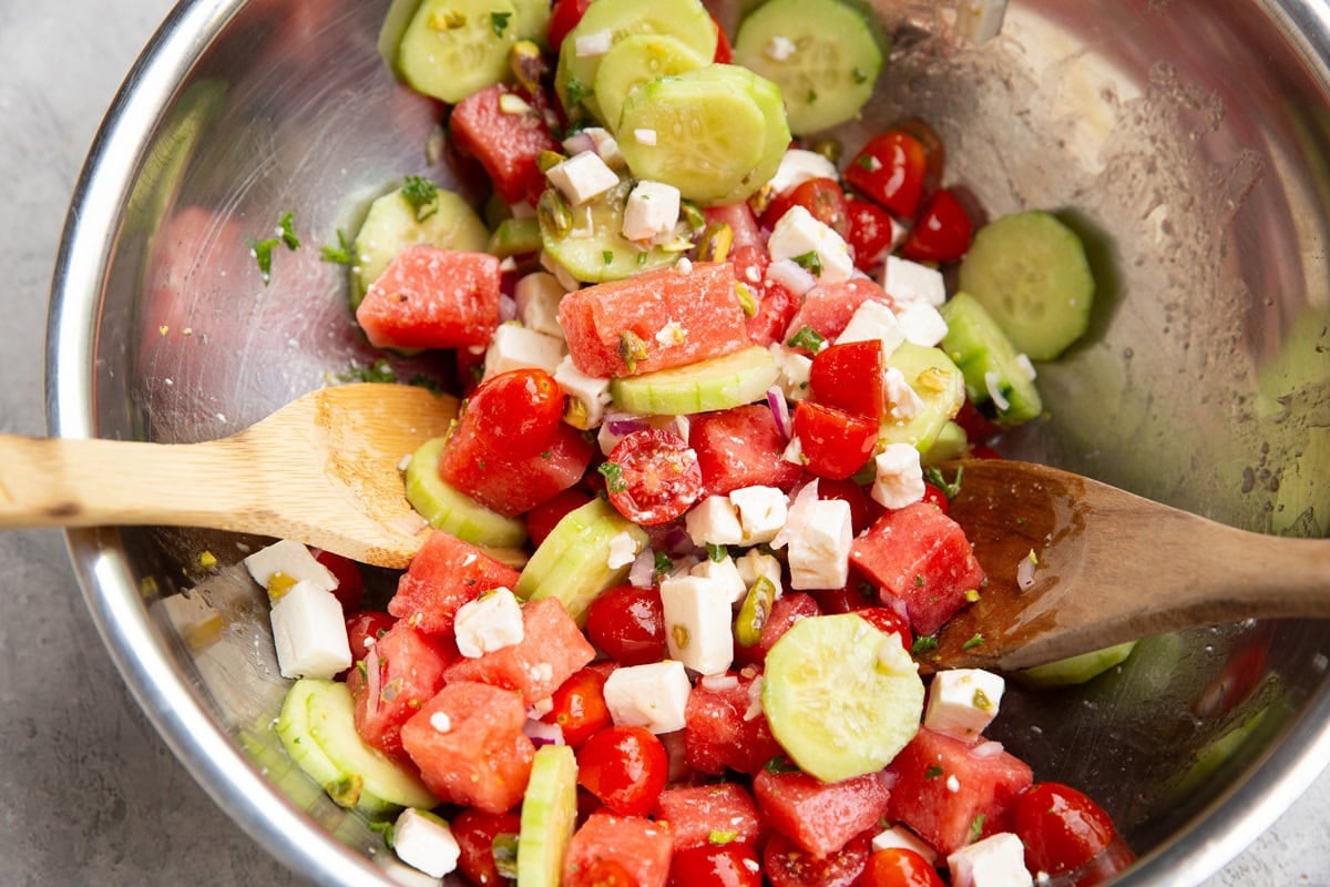 Stainless steel bowl with watermelon feta salad tossed together with two wooden spoons.