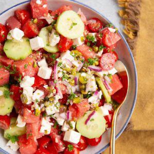 Watermelon salad in a large white blue rimmed bowl with a golden spoon and a golden napkin to the side. The salad includes cucumber, feta cheese, red onion, pistachios and tomatoes in addition to watermelon.