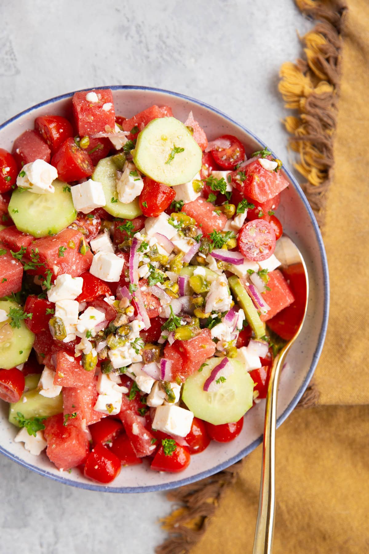 Watermelon salad in a large white blue rimmed bowl with a golden spoon and a golden napkin to the side. The salad includes cucumber, feta cheese, red onion, pistachios and tomatoes in addition to watermelon.